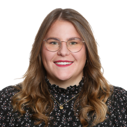 Frau mit langen, braunen Haaren und Brille schaut lächelnd in die Kamera.