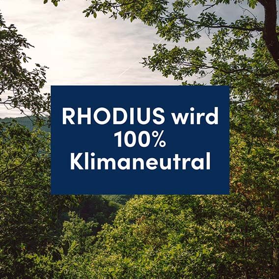 Blick in eine Waldlandschaft. In der Mitte ist eine blaue Fläche zu sehen, auf der in weißer Schrift “RHODIUS wird 100% Klimaneutral” steht.