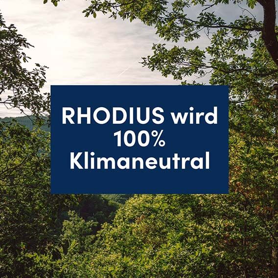 View of a forest landscape. In the centre is a blue area with the words "RHODIUS becomes 100% climate neutral" in white letters.