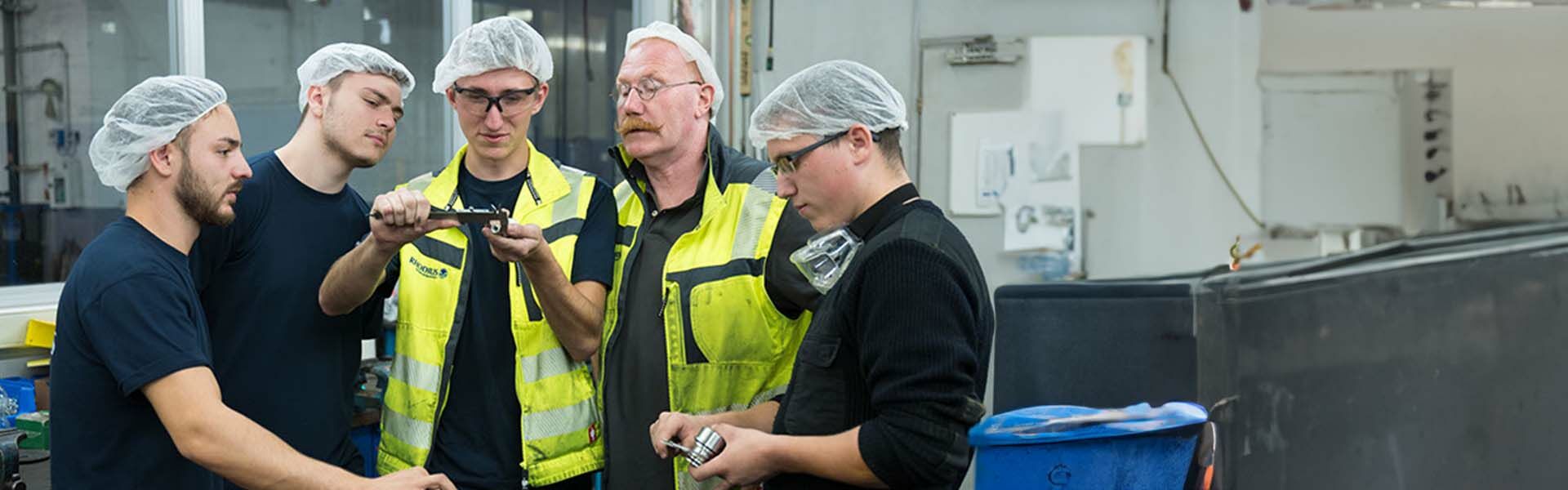 Mann in blauem Arbeitsoverall steht mit vor der Brust verschränkten Armen vor einer Anlage und blickt in die Kamera.